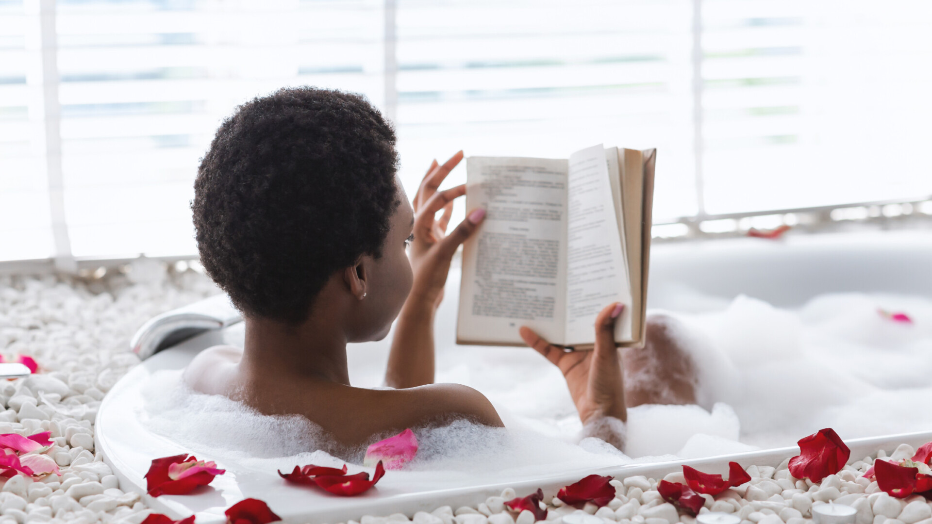 A woman enjoys a relaxing bubble bath on valentine's day self-caring with rose petals, reading a book surrounded by white pebbles and natural light.
